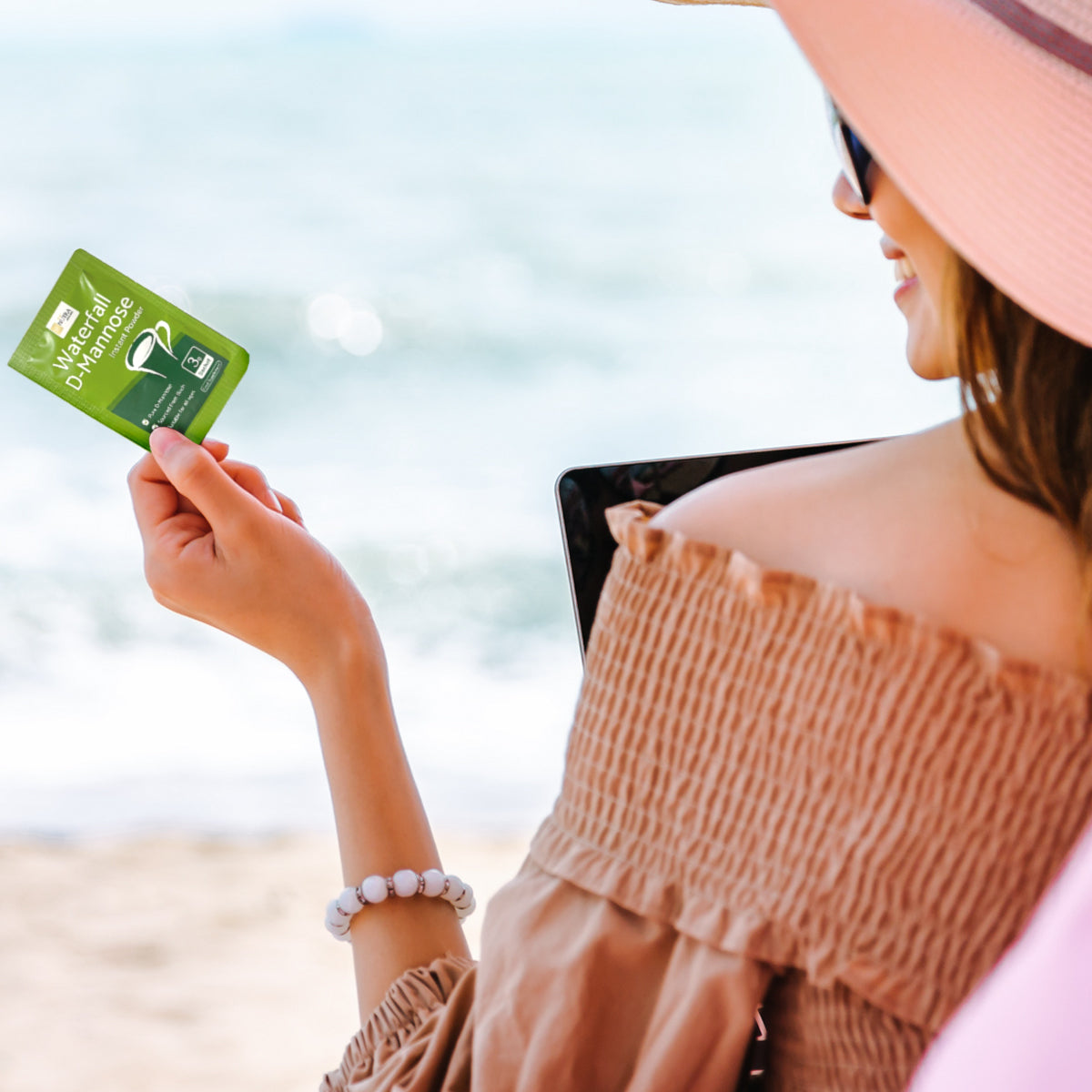 Waterfall D-Mannose sachets lifestyle image with a young woman holding a single sachet in her left hand with the beach in the background – highlighting the sachets suitability for traveling.