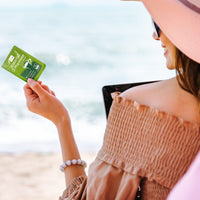 Waterfall D-Mannose sachets lifestyle image with a young woman holding a single sachet in her left hand with the beach in the background – highlighting the sachets suitability for traveling.
